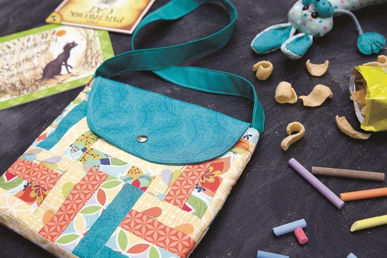 May include: A teal and multi-coloured patchwork fabric crossbody bag with a teal flap and a snap closure. The bag is on a black surface with a toy, crisps, and chalk.