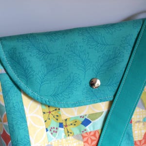 May include: A teal blue crossbody bag with a flap closure and a patterned fabric design. The bag has a teal blue strap and a silver button closure.