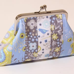 Puede incluir: Un bolso de mano estampado con un cierre plateado. La tela presenta un diseño de patchwork con estampados florales azules, grises y amarillos. Un pequeño pájaro azul aparece en la parte delantera del bolso.