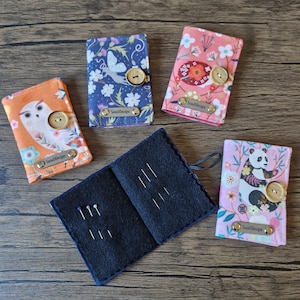 May include: A collection of fabric needle cases with various designs, including an owl, a panda, and floral patterns. Each case has a wooden button closure and a metal tag with the text "SusieDesigns". One case is open, displaying needles.