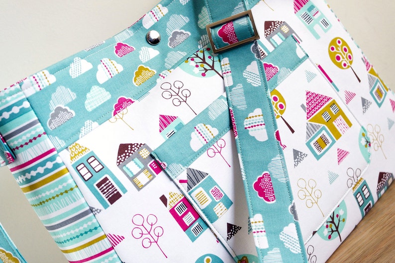 May include: A teal and white fabric tote bag with a colourful house and tree pattern. The bag has a teal strap with a silver buckle.