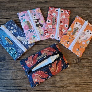 May include: A collection of fabric tissue holders with various patterns, including floral, panda, and owl designs. Each holder has a zipper closure and a small metal tag. The holders are in shades of blue, pink, and orange.