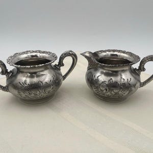 May include: A silver-toned metal sugar bowl with two ornate handles and a matching creamer pitcher with a single handle. Both pieces feature a rounded body, a scalloped rim, and an engraved floral pattern around the exterior surface.