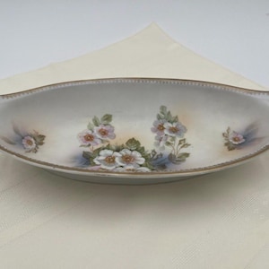 May include: An oval porcelain serving dish with two integrated handles. The white surface features a hand-painted floral motif of pink and white blossoms with green leaves. The rim is decorated with a thin, textured gold-toned metallic border.