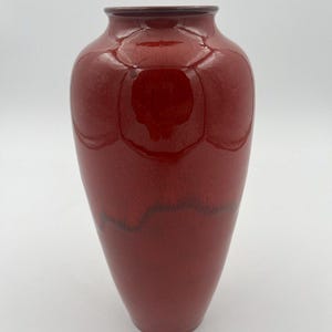 May include: A glossy, deep red ceramic vase with a smooth, rounded body and a narrow, circular opening at the top. The surface features a reflective finish with subtle variations in the red glaze, creating a rich, uniform color throughout the piece.