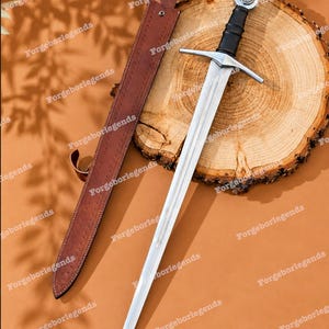 May include: A steel sword with a black grip, a silver crossguard, and a circular pommel, accompanied by a brown leather scabbard with visible stitching and metal rivets. The sword is positioned diagonally next to the scabbard.