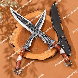 Puede incluir: Dos cuchillos cruzados con hojas de metal martillado curvadas que presentan un patrón oscuro similar a escamas. Los mangos son de madera pulida con detalles en metal plateado y un pomo circular. Una funda de cuero negro se encuentra al lado.