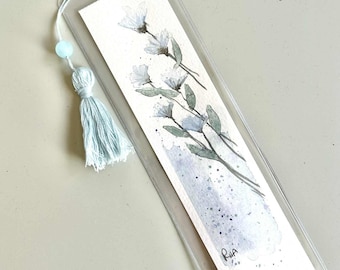 Pode incluir: Marcador de livros em aquarela com um design floral em tons de azul e verde. O marcador de livros é envolto em plástico transparente e tem uma borla e uma conta azuis combinando. As iniciais do artista, "RHA", são visíveis.
