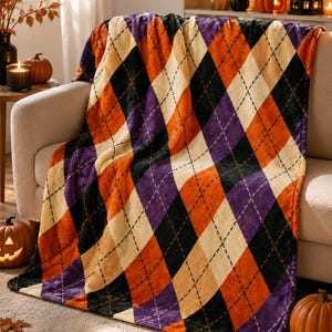 Op de afbeelding: Een zachte plaid met een herhalend argyle ruitpatroon in afwisselende kleuren oranje, paars, zwart en crème. Het ontwerp bevat dunne, gestippelde zwarte lijnen die een raster vormen over de ruitvormen op de stof.