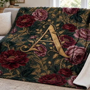 May include: A plush throw blanket featuring a dark green background with a pattern of deep red and pink peonies, gold foliage, and a large, ornate gold letter A centered on the fabric, finished with a white sherpa fleece lining.