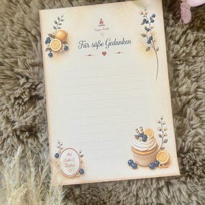 Könnte beinhalten: Ein rechteckiger Notizblock aus cremefarbenem Papier mit horizontalen Linien. Oben steht der Text Sugar Rocks und Für süße Gedanken. Die Illustrationen zeigen Blaubeeren, Zitronenscheiben und einen Cupcake mit Sahnehäubchen.