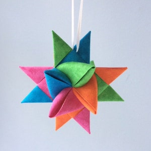 May include: A colorful felt star ornament with a white string for hanging. The star is made of six points, each a different color: pink, orange, green, blue, and two shades of pink.