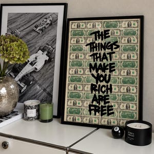 Puede incluir: Una lámina artística enmarcada que muestra una cuadrícula de billetes de un dólar con el texto negro THE THINGS THAT MAKE YOU RICH ARE FREE superpuesto en una fuente negrita y goteante. La lámina está en un marco negro fino sobre una superficie blanca.