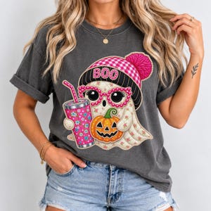 May include: A charcoal gray t-shirt featuring a graphic of a ghost wearing a pink and black plaid beanie with the word BOO, pink glasses, and holding a floral patterned tumbler and a small orange jack-o'-lantern pumpkin with a green stem.