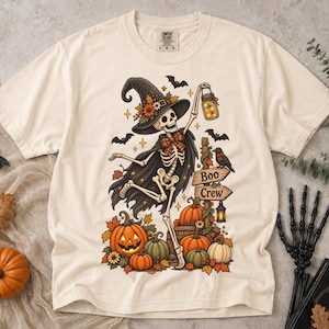 May include: A cream-colored t-shirt featuring a graphic of a skeleton wearing a witch hat and cape, holding a glowing lantern. The design includes pumpkins, bats, a crow, and a wooden signpost with the text Boo Crew, all in autumn colors.