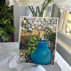 May include: An abstract painting of a blue vase with green leaves and white flowers. The painting is displayed on a white easel. White flowers are in a glass vase in the background. The painting is on a white surface.