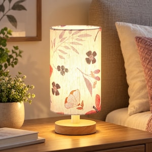 Puede incluir: Una lámpara de mesa cilíndrica con base de madera clara y pantalla de tela con un estampado de elementos botánicos en rojo, rosa y gris oscuro, incluyendo hojas y flores, iluminada desde el interior para emitir una luz cálida.
