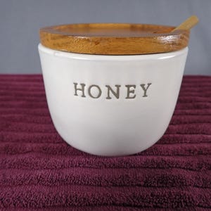 May include: A white ceramic honey pot with a smooth, rounded body and the word HONEY embossed in grey lettering on the front. The container features a circular wooden lid with a small notch and a wooden honey dipper resting inside the pot.