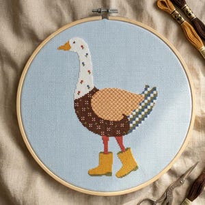 May include: A cross-stitch embroidery project in a round wooden hoop featuring a white goose with a floral neck pattern, a brown patterned body, and yellow rain boots on a light blue fabric background, accompanied by embroidery floss and scissors.