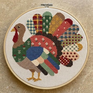 May include: A circular wooden embroidery hoop containing a cross-stitch design of a turkey. The turkey features a brown head and neck with a red wattle, and a body and tail composed of various colorful fabric-like patterns including plaid, floral, and polka dots in shades of red, green, blue, yellow, and brown on a white fabric background.