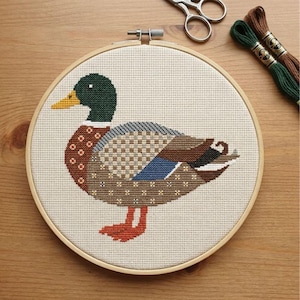 May include: A cross-stitch embroidery of a mallard duck featuring a dark green head, yellow beak, and a body composed of brown, tan, blue, and white geometric patterns, secured within a circular wooden hoop with metal hardware at the top.