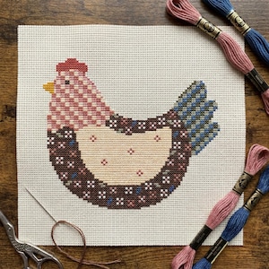 May include: A cross-stitch project on white Aida fabric featuring a chicken with a pink checkered head, a cream-colored body with small pink dots, and a dark brown wing with blue and pink floral accents. Includes embroidery floss and scissors.