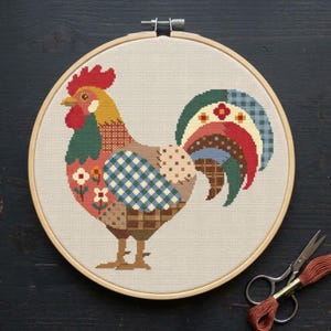 Puede incluir: Un bastidor de bordado circular de madera que contiene un patrón de punto de cruz de un gallo. El gallo presenta una cresta roja, una cabeza amarilla y un cuerpo compuesto por varios patrones geométricos en tonos verde, azul, marrón y rojo.