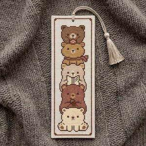 May include: A rectangular cross-stitch bookmark featuring a vertical stack of five stylized bears in shades of brown, tan, and cream. The bookmark has a cream-colored fabric base with a brown border and a beige tassel attached to the top loop.