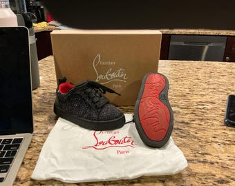 May include: A pair of black glitter trainers with black laces and a red interior lining. One trainer is upright, while the other is positioned to show the signature red sole. The items are displayed with a tan cardboard box and a white dust bag.