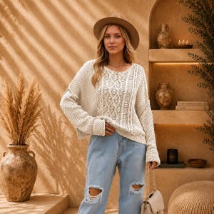 May include: A cream-coloured long-sleeve knit jumper featuring a central cable knit pattern with textured bobbles, paired with light blue distressed denim jeans and a wide-brimmed tan felt hat. The jumper has a relaxed fit and ribbed cuffs.