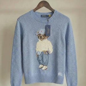Può includere: Un maglione girocollo azzurro con un motivo lavorato a maglia di un orsacchiotto che indossa un berretto bianco, un maglione color crema e jeans blu. Il maglione presenta collo, polsini e orlo a coste, con le lettere RL ricamate in bianco.