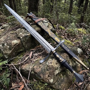 May include: A long sword with a patterned Damascus steel blade, a black leather-wrapped handle with gold-coloured accents, and a matching black scabbard featuring intricate gold-coloured filigree and a brown leather strap for carrying.