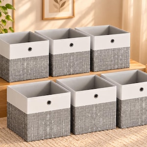 Puede incluir: Un conjunto de seis cajas de almacenamiento cuadradas con un diseño bicolor, con una base de tela gris texturizada y una sección superior blanca sólida. Cada caja incluye un ojal metálico plateado centrado en el panel frontal y un interior gris.