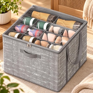 May include: A square, gray storage bin with a textured fabric finish and a clear, zippered top lid. The interior is divided into multiple compartments, each containing various rolled garments and small items in shades of tan, green, and pink.
