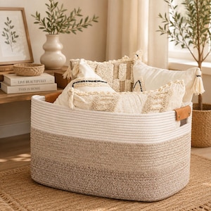 May include: A large, oval-shaped storage basket made of woven cotton rope, featuring a two-tone design with a white upper section and a tan lower section. The basket includes two brown leather-like side handles and is filled with decorative pillows.