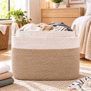 May include: A large, oval-shaped storage basket constructed from coiled rope. The basket features a two-tone design with a natural tan bottom section and a cream-colored top section. It includes two integrated rope handles on the sides.
