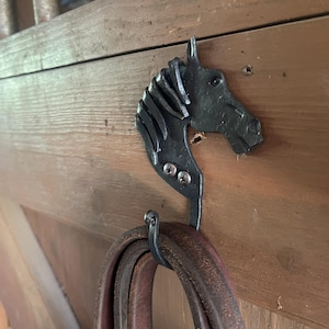 May include: A dark gray metal wall hook shaped like a horse head with a flowing mane. The hook is secured to a wooden surface with two visible silver screws and holds a coiled brown leather strap hanging from its lower curved metal section.