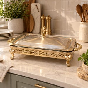 May include: A rectangular gold-toned metal serving dish with a matching lid, featuring an ornate filigree side pattern, decorative handles, and four claw-style feet. A central finial sits atop the lid, providing a handle for lifting.