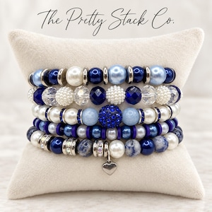 May include: A stack of five beaded bracelets featuring a mix of blue, white, and silver-toned beads. The bracelets include pearl-like, faceted, and smooth round beads, with a small silver heart charm hanging from the bottom strand. Text reads The Pretty Stack Co.