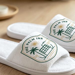 May include: A pair of white open-toe slippers featuring a printed logo that reads The Palm Inn. The logo includes a green palm tree, a sun, and a building outline enclosed within a shield-shaped border with gold and green line accents.