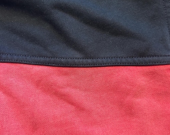 May include: A textile garment featuring a horizontal seam that divides the fabric into two distinct sections. The upper portion is a solid dark black color, while the lower portion is a solid vibrant red color. The fabric has a visible knit texture.