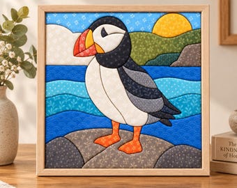 May include: A square framed fabric art piece featuring a quilted puffin standing on a gray rock. The bird has a black and white body with a bright orange beak and feet. The background shows a blue sea, green hills, and a yellow sun.