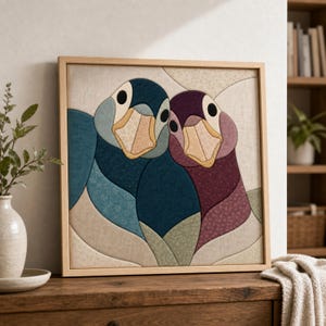 Pode incluir: Uma peça de arte de parede quadrada emoldurada com um design de patchwork de tecido de dois gansos lado a lado. O ganso da esquerda é representado em tons de azul e azul-petróleo, enquanto o da direita é em tons de ameixa e roxo.