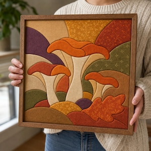 Può includere: Un'opera d'arte murale quadrata in tessuto all'interno di una cornice in legno, caratterizzata da un design trapuntato a strati con tre funghi nei toni dell'arancione, crema e marrone, su uno sfondo di toppe in tessuto verde, viola e oro.