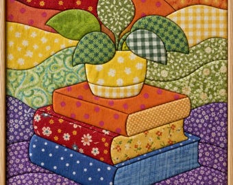 May include: A framed fabric wall art piece featuring a stack of three books in orange, red, and blue with floral and polka dot patterns. A yellow pot with a green leafy plant sits on top. The background consists of wavy, multicolored fabric layers.