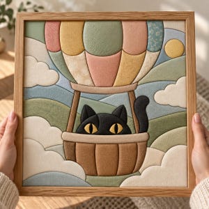 May include: A square framed fabric wall art piece featuring a black cat with yellow eyes sitting in a hot air balloon. The balloon has panels of muted pink, yellow, green, and blue fabric. The background shows rolling hills and clouds.