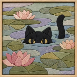 May include: A framed fabric wall art piece featuring a black cat with yellow eyes peeking out of a pond filled with green lily pads and pink lotus flowers. The scene is constructed from layered fabric segments in shades of green, blue, and pink.