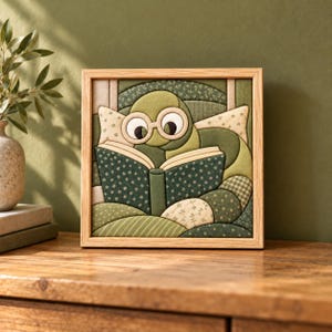 May include: A square framed fabric wall art piece featuring a green worm wearing round glasses and reading an open book. The design is composed of various green and cream fabric patches with floral, striped, and checkered patterns.