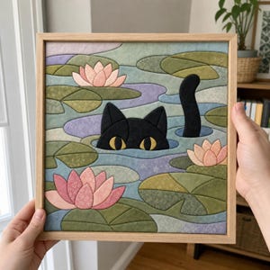 May include: A square framed fabric art piece featuring a black cat with yellow eyes peeking out of a pond filled with green lily pads and pink lotus flowers. The scene is constructed from various patterned fabric segments in a mosaic style.