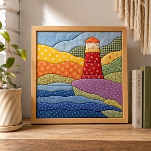 May include: A square fabric wall art piece in a light wood frame featuring a quilted landscape scene. The design includes a red lighthouse with floral patterns, rolling hills in yellow, orange, and green, and blue wavy water sections.